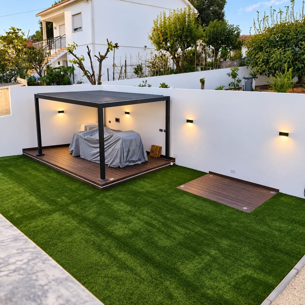 Quintal renovado com relva sintética, deck em madeira compósita, pérgola e apliques de luz nos muros pintados de branco.