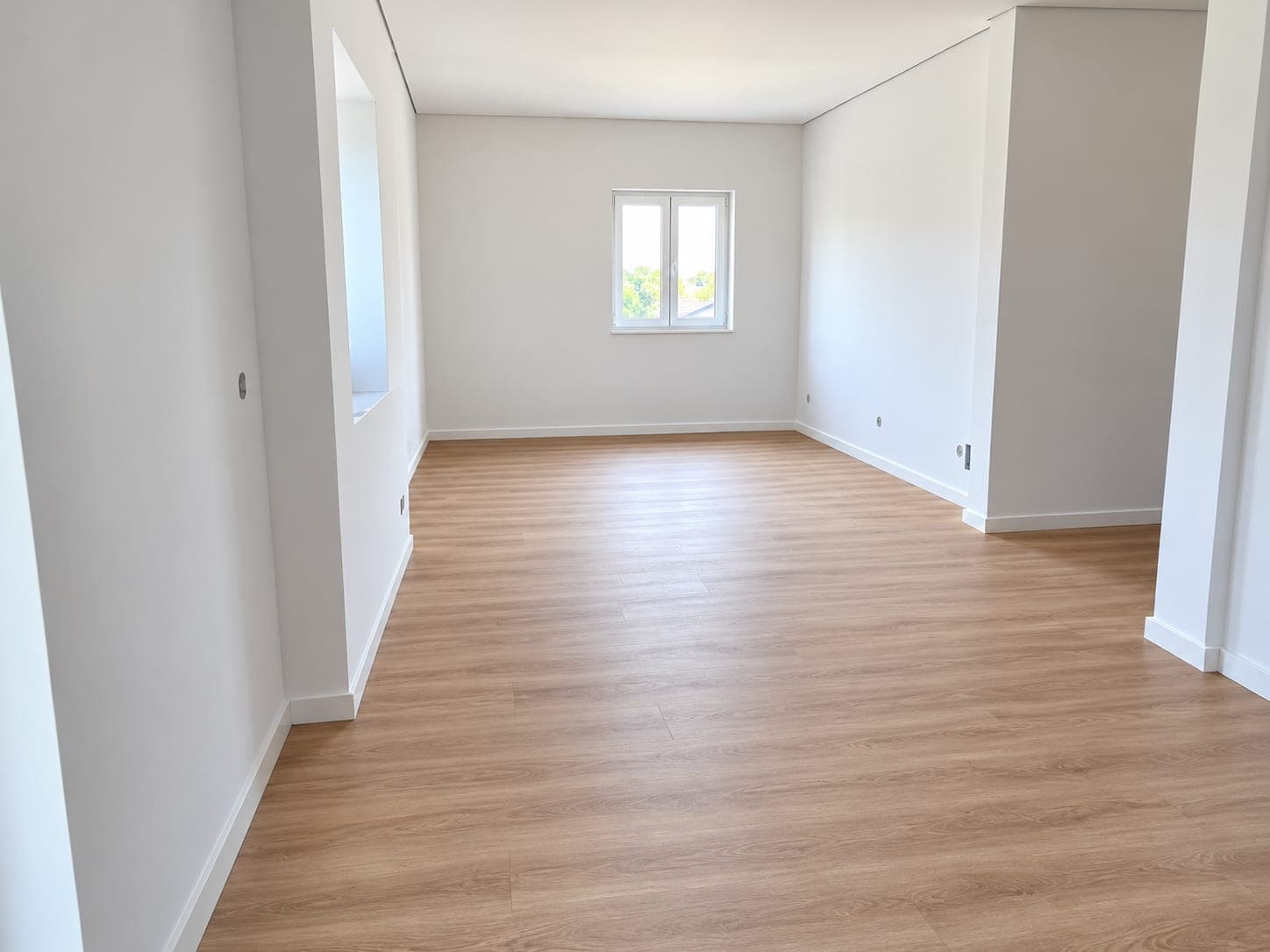 Sala ampla e luminosa com paredes brancas, rodapés novos e pavimento em madeira clara.